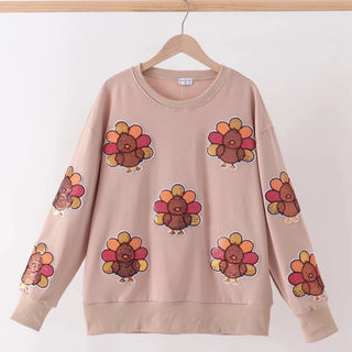 A beige crewneck sweatshirt with a repeating pattern of cartoon turkeys in autumn colors, hanging on a wooden hanger against a plain white wall.