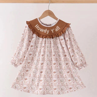 A long-sleeve dress with a brown ruffled collar printed with a light pink and white circular pattern, hanging on a wooden hanger against a plain white wall. The collar is embroidered with the text 'Howdy Y'all'.