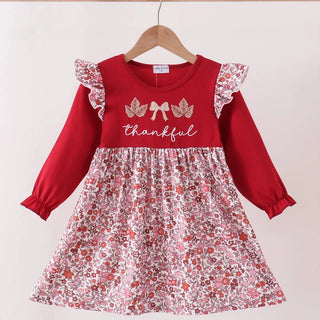 A toddler girl's long-sleeve dress hanging on a wooden hanger against a plain light background. The dress has a solid red bodice with 'thankful' and a bow design on the chest, and a red, white, and pink floral print skirt with ruffled sleeves.