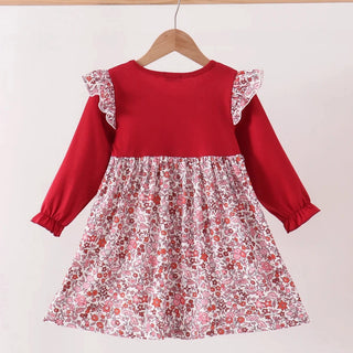 A toddler girl's long-sleeve dress hanging on a wooden hanger against a plain white wall. The dress features a solid red yoke and sleeves with ruffle details, and a gathered skirt in a pink, red, and white floral pattern.