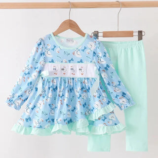 A two-piece baby girl's outfit set hanging on wooden hangers against a plain white wall. The set includes a light blue long-sleeve ruffled dress with a white smocked chest panel and goose print, paired with mint green leggings.