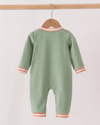 A sage green baby jumpsuit hanging on a wooden hanger from a light wood rod, featuring orange and white striped trim at the collar, cuffs, and cuffs.