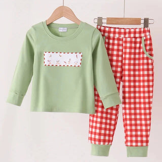 A two-piece children's outfit set hanging on a wooden rod. The set includes a light sage green long-sleeve top with a rectangular chest patch featuring a red and white geese  pattern, and matching red and white gingham checkered pants with sage green cuffs.