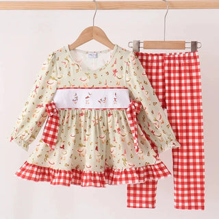 A toddler girl's two-piece clothing set hanging on wooden hangers against a plain white wall. The set includes a long-sleeve top with a cream base, a red and white cherry print, a white smocked chest panel with goose motifs, and red and white gingham ruffle trim, paired with matching red and white gingham pants.
