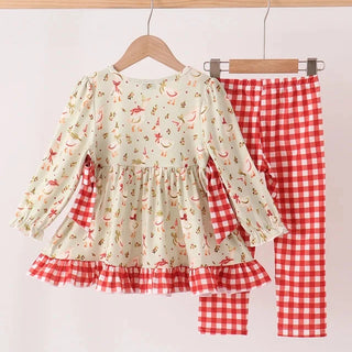 A two-piece children's outfit hanging on wooden hangers against a plain light background. The set includes a long-sleeve cream-colored top with a small red and green holiday print, red-and-white gingham ruffle trim at the hem and cuffs, and matching red-and-white gingham pants.