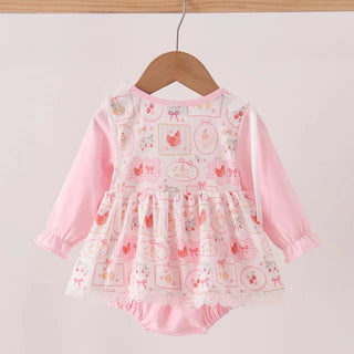 A baby girl's long-sleeve romper dress in light pink, hanging on a wooden hanger. The dress features a solid pink bodice and sleeves, with a tiered skirt overlay printed with a white grid pattern of small pink chickens, eggs, and bows, finished with a white lace hem.