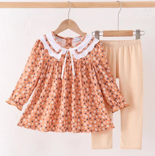 A toddler girl's two-piece outfit hanging on wooden hangers against a plain white wall. The set includes a peach-orange long-sleeve blouse with a white floral print, white ruffled Peter Pan collar, and white tie detail, paired with solid beige leggings.