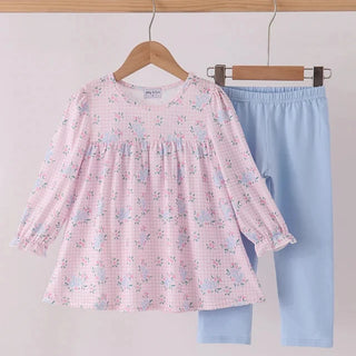 A toddler girl's two-piece clothing set hanging on wooden hangers against a light gray wall. The set includes a long-sleeve smocked blouse in light pink with a small blue and white floral pattern, and matching light blue leggings.