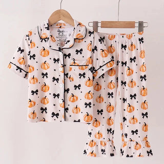 A two-piece children's pajama set hanging on wooden hangers against a plain light background. The set includes a short-sleeve button-front top and matching pants, both in a warm off-white fabric with an allover print of orange pumpkins and black bows.