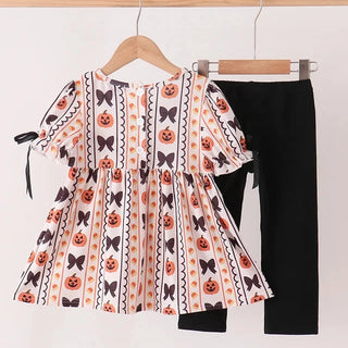 A toddler girl's two-piece Halloween outfit hanging on wooden hangers against a plain white wall. The set includes a short-sleeve peplum top with an orange, black, and white pumpkin and bow pattern, paired with solid black pants.