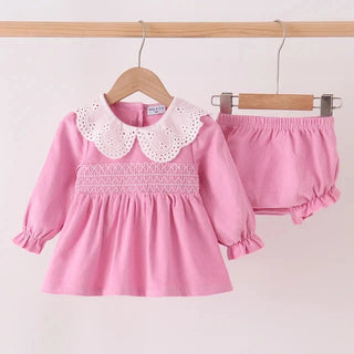 A pink toddler girl's two-piece outfit set consisting of a long-sleeve dress with a white eyelet Peter Pan collar and smocked chest detail, and matching pink ruffled bloomers, hanging on wooden hangers against a plain light background.