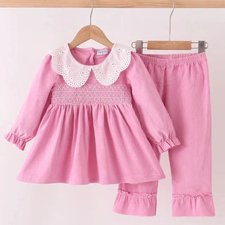 A two-piece toddler girl's outfit set in pink, featuring a long-sleeve smocked dress with a white eyelet Peter Pan collar and matching pink ruffled pants, displayed on wooden hangers against a white background.