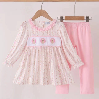 A toddler girl's two-piece clothing set hanging on wooden hangers against a light pink background. The set includes a long-sleeve dress with a white base, pink floral print, and a pink ruffled collar, paired with solid light pink pants.
