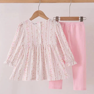 A two-piece children's outfit hanging on wooden hangers against a plain light background. The set includes a long-sleeve ruffled blouse in a white fabric with a delicate pink floral pattern, paired with solid light pink pants.