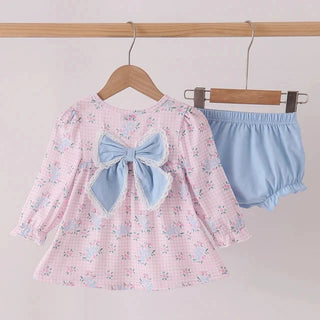 A toddler girl's two-piece outfit hanging on wooden hangers from a wooden rod. The set includes a light pink gingham long-sleeve top with a large light blue bow on the back, and matching light blue bloomers.