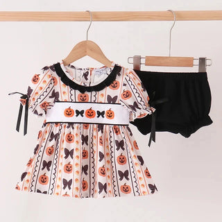 A toddler girl's Halloween outfit set hanging on wooden hangers against a plain white wall. The set includes a short-sleeve dress with a white base, black trim, and an all-over print of orange pumpkins, black bows, and black-and-white zigzag stripes, paired with black ruffled bloomers.