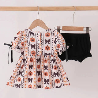 A toddler girl's Halloween outfit set hanging on a wooden rod, featuring a short puff-sleeve dress with an orange, black, and white pumpkin and bow pattern, and a pair of black ruffled shorts.