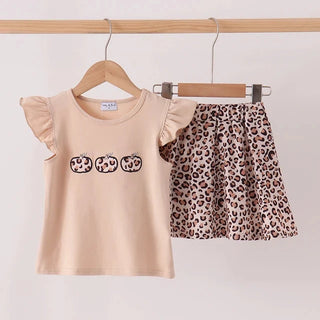 A two-piece girls' outfit hanging on wooden hangers against a plain light background. The set includes a light beige short-sleeve top with ruffled sleeves and three small leopard-print patch details on the chest, paired with a leopard-print skirt.