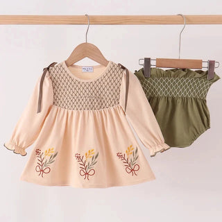 A two-piece baby girl's outfit hanging on a wooden hanger, featuring a long-sleeve peach dress with a beige geometric patterned yoke and embroidered floral details, paired with an olive green smocked bloomers set, displayed against a plain light background.