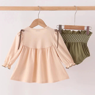 A toddler girl's two-piece clothing set hanging on wooden hangers from a wooden rod. The set includes a long-sleeve peplum top in a light peach-beige color with dark brown ribbon ties at the shoulders, and matching olive green smocked bloomers with a white lattice pattern.