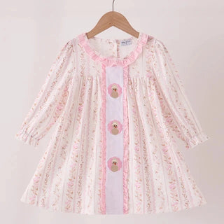 A toddler girl's long-sleeve dress with a white base, light pink floral print, and a central pink lace-trimmed placket with three decorative buttons, hanging on a wooden hanger.
