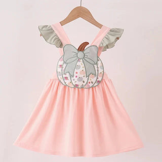A girl's dress hanging on a wooden hanger against a light pink background. The dress is light pink with ruffled sleeves and features a decorative front applique shaped like a pumpkin with a gray bow and floral pattern.
