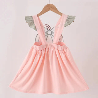 A light pink sleeveless dress with ruffled shoulder details and a gathered skirt, hanging on a wooden hanger against a plain light background.