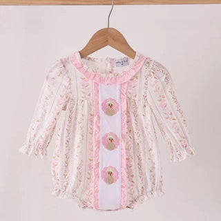 A baby girl's long-sleeve romper in white and pink floral lace, hanging on a wooden hanger against a plain light beige wall. The romper features a white center panel with pink ruffles, three decorative bear-shaped buttons, and a pink collar.