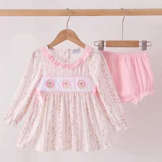 A baby girl's two-piece clothing set hanging on wooden hangers against a light pink background. The set includes a long-sleeve smocked dress in white with a delicate pink floral print, ruffled collar, and a pink smocked chest panel with three embroidered turkeys, paired with matching pink ruffled bloomers.