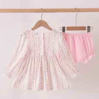 A two-piece baby girl's outfit set hanging on wooden hangers from a wooden rod. The set includes a long-sleeve white tunic top with a delicate pink and green floral pattern, and a pair of light pink bloomers.