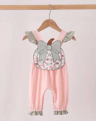 A baby romper in light pink with a decorative applique of a gray bow and a patterned pumpkin on the chest, hanging on a wooden hanger against a light pink background.