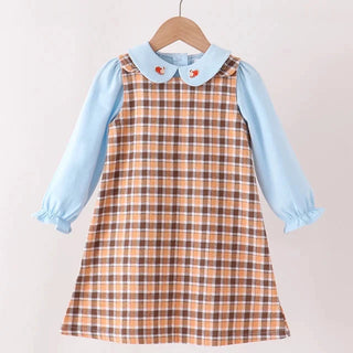 A toddler girl's dress hanging on a wooden hanger against a light beige wall. The dress features a light blue long-sleeve shirt with ruffled cuffs and collar, layered under a brown and beige plaid pinafore-style dress with a matching collar.