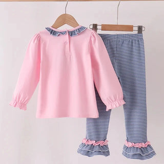 A two-piece children's outfit hanging on wooden hangers from a wooden rod. The set includes a light pink long-sleeve top with a blue and white patterned ruffled collar, and matching blue-and-white striped pants with pink ruffled cuffs.