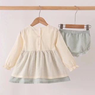 A baby girl's two-piece outfit set consisting of a cream-colored long-sleeve dress with a green and white gingham hemline, and matching green and white gingham bloomers, hanging on wooden hangers from a wooden clothes rack.