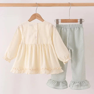 A two-piece baby clothing set hanging on a wooden hanger bar. The set includes a cream-colored long-sleeve blouse with a ruffled hem and matching green-and-white gingham check pants with ruffled hems.