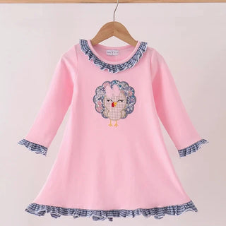 A pink long-sleeve dress with blue and white gingham ruffle trim on the collar, cuffs, and hem, featuring a central embroidered turkey design, hanging on a wooden hanger.