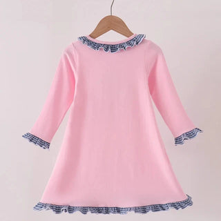A pink long-sleeve dress with blue and white patterned ruffle trim on the collar, cuffs, and hem, hanging on a wooden hanger against a light pink background.