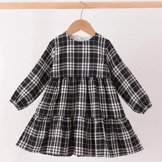 A long-sleeve tiered dress for toddlers, featuring a black and white plaid pattern, hanging on a wooden hanger from a light wood clothing rack against a plain white background.