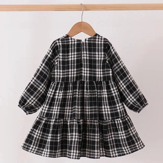 A black and white plaid tiered dress with long sleeves hanging on a wooden hanger from a light wood clothes rack against a plain white wall.