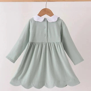 A pale sage green long-sleeve baby dress with a white Peter Pan collar and scalloped hem, hanging on a wooden hanger against a light beige background.