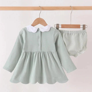 A baby girl's two-piece outfit set consisting of a sage green long-sleeve dress with a white Peter Pan collar and matching green-and-white checkered bloomers, hanging on wooden hangers from a light wood clothing rack against a plain off-white wall.