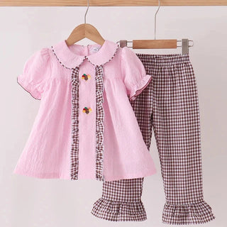 A toddler girl's two-piece clothing set hanging on wooden hangers against a plain light background. The set includes a light pink short-sleeve blouse with a Peter Pan collar, ruffle trim, and decorative turkeys, paired with brown and white gingham check pants featuring ruffled hems.