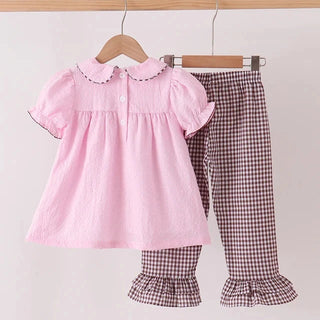 A toddler girl's two-piece clothing set hanging on wooden hangers from a light wood rod. The set includes a light pink short-sleeve blouse with a ruffled Peter Pan collar and matching brown and white gingham pants with ruffled hems.