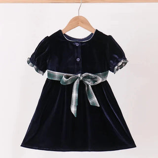 A toddler girl's navy blue velvet dress with short puff sleeves, a buttoned neckline, and a green and white plaid ribbon bow at the waist, hanging on a wooden hanger against a white wall.