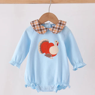 A baby long-sleeve romper in light blue, hanging on a wooden hanger. The romper features a plaid collar in brown, orange, and white, and a central applique of a turkey.