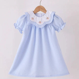 A light blue girl’s dress with a white ruffled Peter Pan collar decorated with small embroidered turkeys, hanging on a wooden hanger from a wooden rod against a plain light background.
