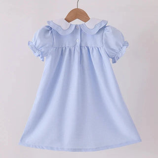 A light blue girl’s dress with a white Peter Pan collar and ruffled short sleeves, hanging on a wooden hanger from a wooden clothes rack against a plain light background.