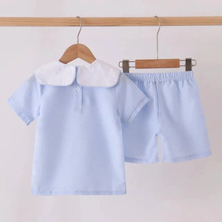 A light blue children's short-sleeve top with a white Peter Pan collar, shown hanging on a wooden hanger next to matching light blue shorts on a wooden hanger, suspended from a wooden rod against a plain light background.