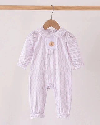 A baby romper in a very light pink and white micro-dot pattern, hanging on a wooden hanger from a wooden clothes rack. The romper features a Peter Pan collar, ruffled cuffs, and ruffled hems, with a small embroidered turkey on the chest.