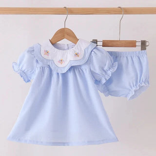 A baby girl's two-piece outfit set consisting of a light blue short-sleeve dress with a white Peter Pan collar decorated with small turkey embroidery, and matching light blue bubble shorts, hanging on wooden hangers against a plain off-white wall.